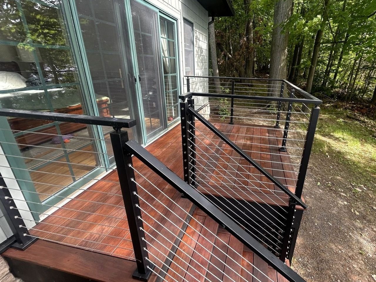 Hardwood deck and steps with black steel posts and stainless cable railing beside a shingled house in the woods